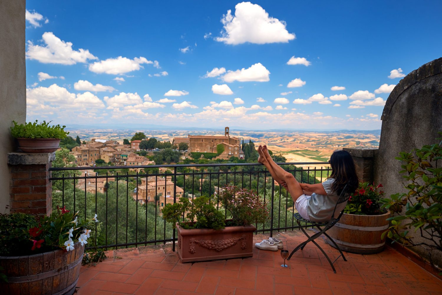 Summer Holidays in Tuscany. A middle-aged woman with long hair sitting in an armchair with her legs resting on a railing, watching city landscape from a terrace of a stone house. Montalcino, Italy.